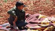 A child sits next to an old woman lying on a blanket on the ground.