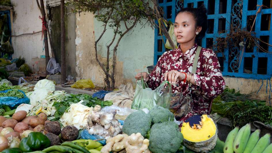 Eine Verkäuferin aus Kambodscha steht hinter einem prall gefülltem Tisch mit Gemüse.