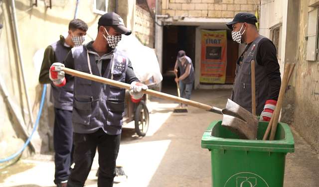 Drei Männer bei der Müllbeseitigung: Ein Mann wirft mit einer großen Schaufel etwas in eine grüne Tonne.