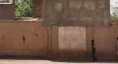 Child standing in front of a house in Sudan after the outbreak of war, smoke rising.