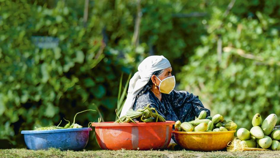 Eine Frau mit Mundschutz und einem Tuch um den Kopf sitzt auf dem Boden, neben sich stehen Schüsseln mit Gemüse.