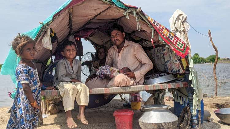 Fluten in Pakistan: Ein Mann, eine Frau, zwei kleine Kinder und ein Baby in ihrer Notunterkunft, die aus Holzstäben, Decken und einer Plastikabdeckung besteht. im Hintergrund ist Wasser zu sehen.