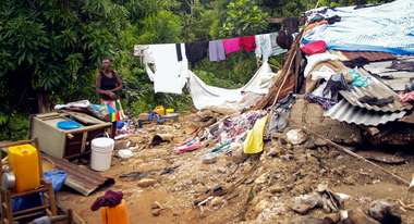 Eine Frau steht inmitten von Schutt und Teilen eines zerstörten Hauses. Im Hintergrund ist Wäsche aufgehängt. 