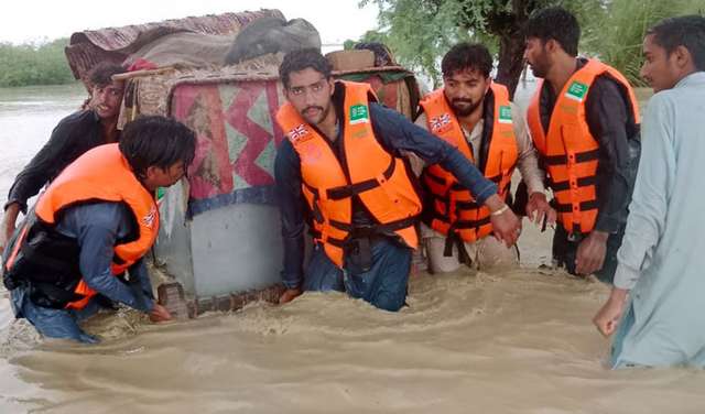 Überschwemmungen in Pakistan. Männer mit Schwimmwesten tragen einen großen Container durch kniehohes Wasser