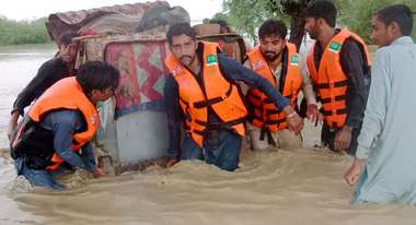 Überschwemmungen in Pakistan. Männer mit Schwimmwesten tragen einen großen Container durch kniehohes Wasser