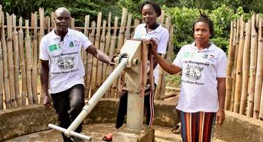 Two women and a man stand next to a water pump and look into the camera.