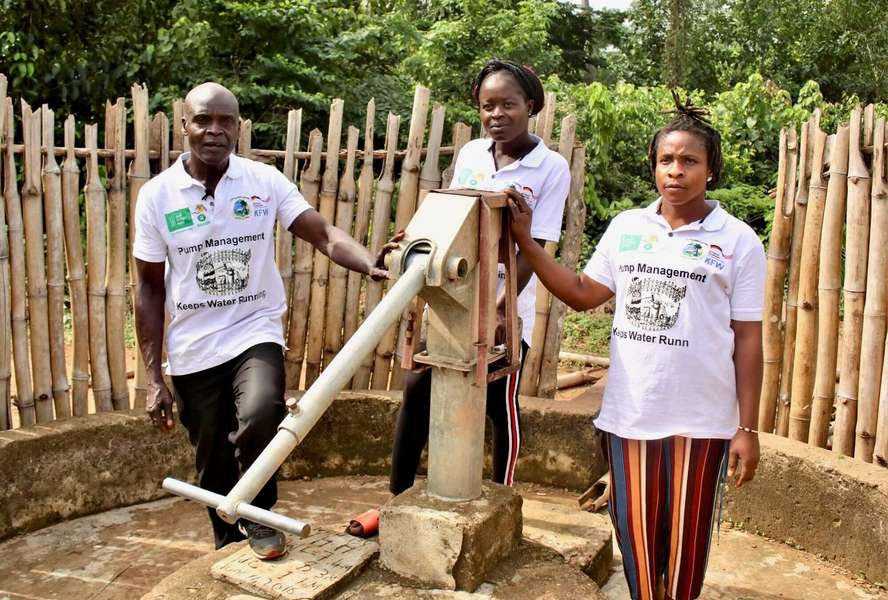 Two women and a man stand next to a water pump and look into the camera.