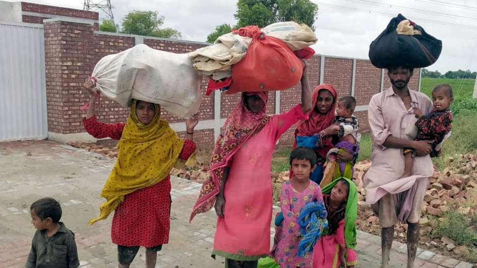 Überschwemmungen in Pakistan. Eine neunköpfige Familie in bunter Kleidung trägt ihr weniges Hab und Gut auf den Köpfen. Sie sind barfuß. 