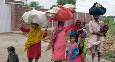 Überschwemmungen in Pakistan. Eine neunköpfige Familie in bunter Kleidung trägt ihr weniges Hab und Gut auf den Köpfen. Sie sind barfuß. 