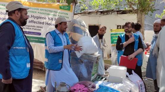 Verteilung von Hygiene-Kits in Pakistan: Männer in blauen Westen unterhalten sich, im Hintergrund ist ein Sack mit warmen Decken zu sehen.