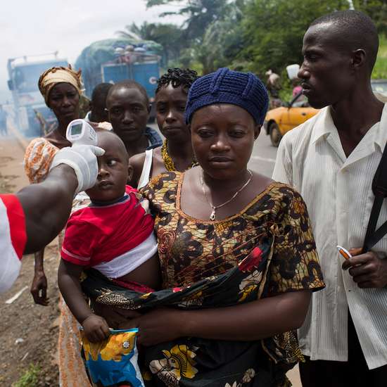 Eine Frau trägt ein Kleinkind auf dem Arm. Ein Mann misst misst mit einem Fieberthermometer die Temperatur des Kindes.