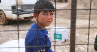 Ein Junge sitzt hinter einem Gitter und schaut in die Kamera. Im Hintergrund steht ein Paket der Welthungerhilfe.