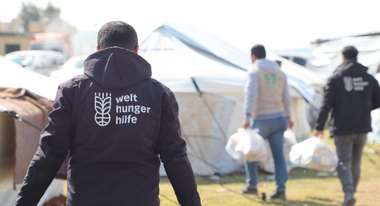 Distribution of hot meals in a refugee camp in Syria. In the foreground, an aid worker with the Welthungerhilfe logo on the back of his jacket.