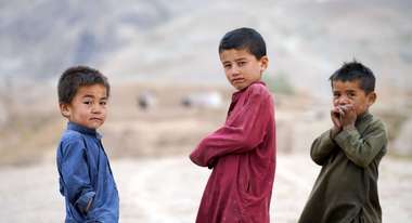 Drei Kinder in Afghanistan schauen in die Kamera. Im Hintergrund ist karge Landschaft zu sehen.