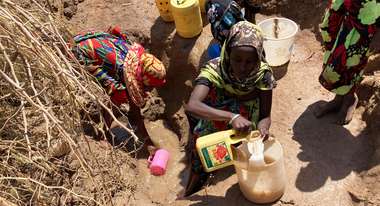 Mehrere Frauen holen mit gelben Kanistern Wasser aus einem Erdloch.