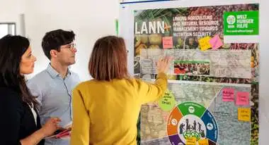 Drei Personen betrachten ein Flipchart und kleben Notizzettel mit Schlagworten darauf.