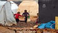 Three children holding rubber boots amongst tents in a refugee camp.