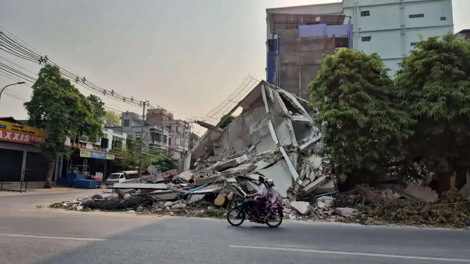 Destroyed bank building in the city of Mandalay Zwei Menschen auf einem Mofaroller fahren an einem zerstörten Bankgebäude an einer Kreuzung vorbei