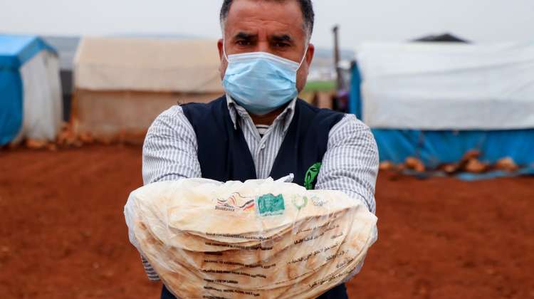 Ein Helfer mit Mundnasenschutz zeigt einen Stapel Fladenbrote in einem Camp für Binnenvertrieben in der syrischen Region Aleppo.