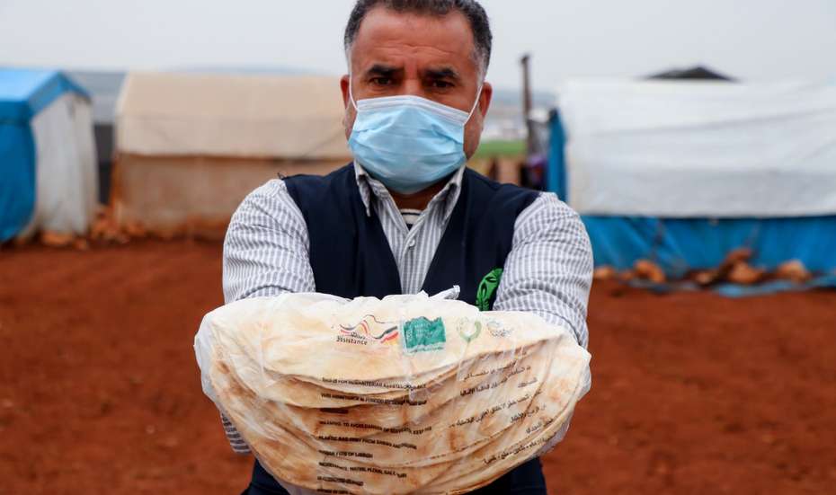 Ein Helfer mit Mundnasenschutz zeigt einen Stapel Fladenbrote in einem Camp für Binnenvertrieben in der syrischen Region Aleppo.