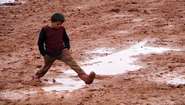 A boy wearing rubber boots walks through mud and puddles